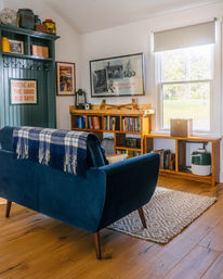 Sunlit cozy living room in a rustic cottage with a blue velvet mid-century sofa draped in a plaid blanket, wooden bookshelves, vintage camp-themed prints and a large window overlooking greenery.