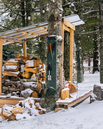 Cozy snowy scene: rustic wood shelter with stacked firewood, outdoor tools and hanging string lights tucked among mossy trees in a winter forest