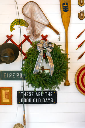 Festive evergreen holiday wreath with silver bow on a white shiplap wall, surrounded by vintage lake-cabin decor — wooden oar, fishing net, hat, arrows and a black sign reading 'THESE ARE THE GOOD OLD DAYS'.