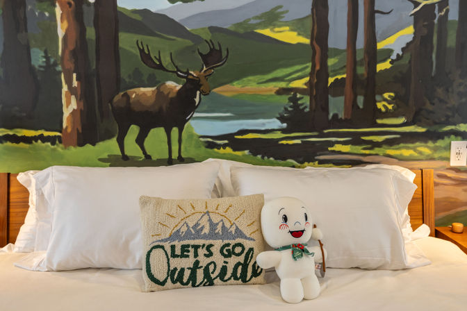 Cozy mountain-cabin bedroom with white bedding, embroidered “Let’s Go Outside” pillow, cheerful plush toy on the bed, and a large mural of an elk by a forested lake