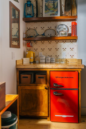 Cozy rustic kitchenette nook with a bright red retro mini fridge, wooden cabinet and counter, open shelves of vintage decor, yellow kettle and labeled coffee, tea and sugar canisters against crossed‑paddle wallpaper.