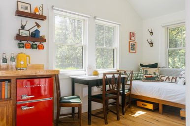 Sunlit cozy cabin-style kitchenette and dining nook with a retro red mini fridge, yellow kettle on a wooden counter, small black table with wooden chairs, daybed with camp-themed pillows, floating shelves with colorful mugs, and large windows looking onto leafy trees.