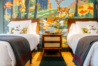 Cozy boutique hotel room with two twin beds, smiling plush toys on pillows, a mid-century nightstand with twin lamps, and a colorful woodland mural featuring a deer and autumn trees.