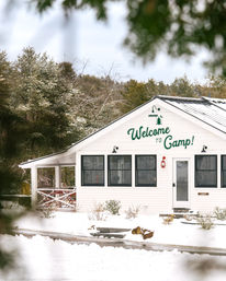 Snow-covered white camp lodge with green "Welcome to Camp!" lettering, black-trim windows and front porch, nestled among evergreen trees in a winter woodland retreat.