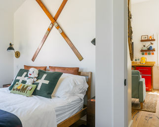 Cozy cabin-style bedroom with crossed vintage wooden skis on a white wall, a bed topped with green camp-themed pillows and a plush toy, brass wall sconce, and a peek through the doorway to a living nook with a red mini fridge and green armchair.