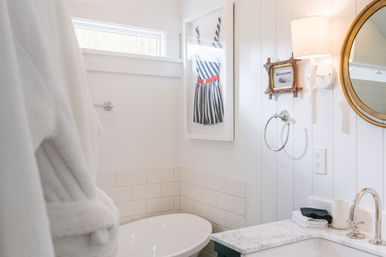 Bright coastal-style white bathroom with a freestanding tub, marble vanity and chrome faucet, round mirror and wall sconce, playful framed striped dress artwork and a plush white robe.