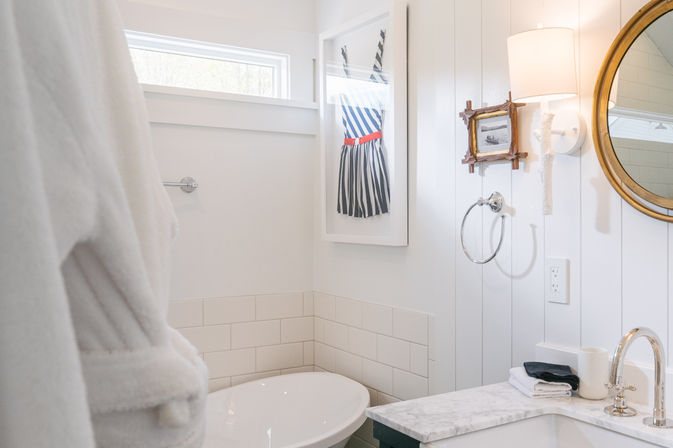 Bright coastal-style white bathroom with a freestanding tub, marble vanity and chrome faucet, round mirror and wall sconce, playful framed striped dress artwork and a plush white robe.