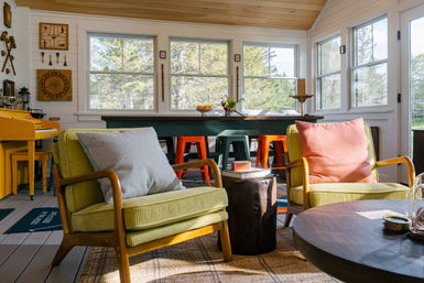 Sunlit cabin-style sunroom with large windows framing a forest view, mid-century green armchairs with patterned pillows, colorful bar stools at a long counter, a yellow piano and a wooden stump side table — cozy, bright retreat.