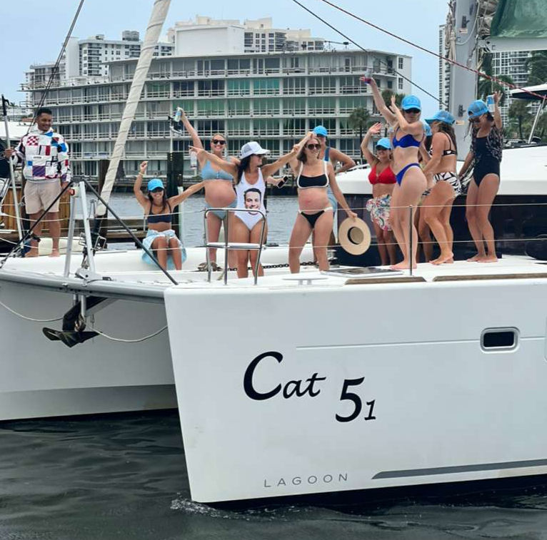 Boat party on a white catamaran — women in swimsuits and blue caps dancing and posing on deck at a marina with waterfront condos in the background.