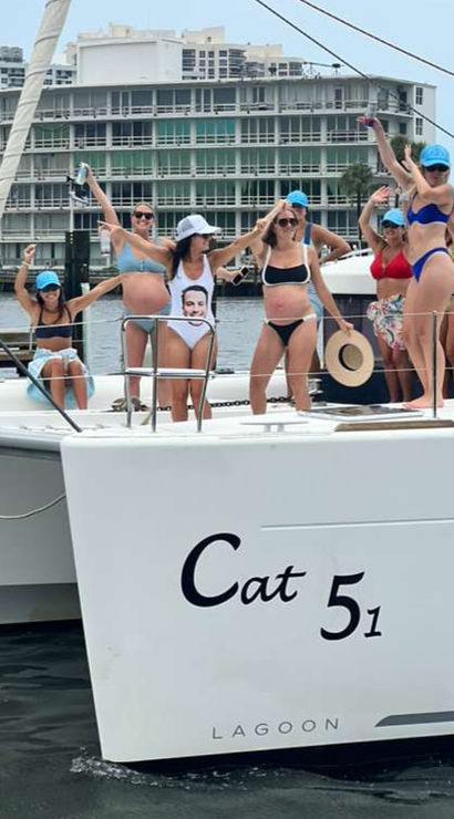 Boat party on a white catamaran — women in swimsuits and blue caps dancing and posing on deck at a marina with waterfront condos in the background.