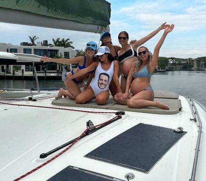 Five women in swimsuits posing and cheering on the bow of a white yacht in a sunny waterfront marina with palm trees and modern homes