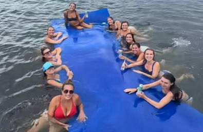 Smiling women in swimsuits lounging along a large blue floating mat in calm water, holding canned drinks — a fun summer lake float with friends.