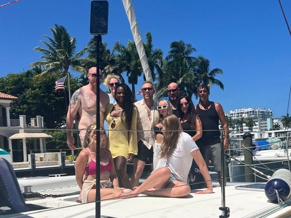 Sun-soaked group of friends posing on a sailboat deck at a palm-lined marina with bright blue sky, docked boats and an American flag.