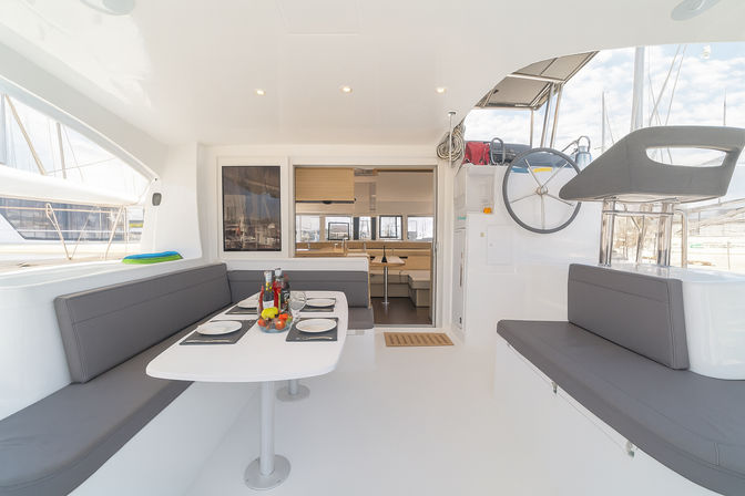 Bright modern yacht cockpit dining area with white surfaces and gray cushioned benches, table set for four with fruit and drinks, helm wheel and captain’s seat, cabin interior visible and sailboat masts at a marina beyond.