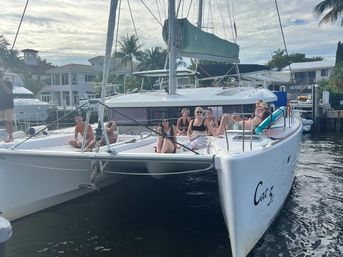 Group of people lounging on the foredeck of a white catamaran at a sunny tropical marina with palm trees, waterfront homes and docked boats