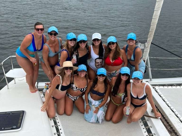 A dozen women in bikinis and matching blue caps smiling and holding drinks while posing on the netted bow of a catamaran over calm gray water — lively summer boat party.