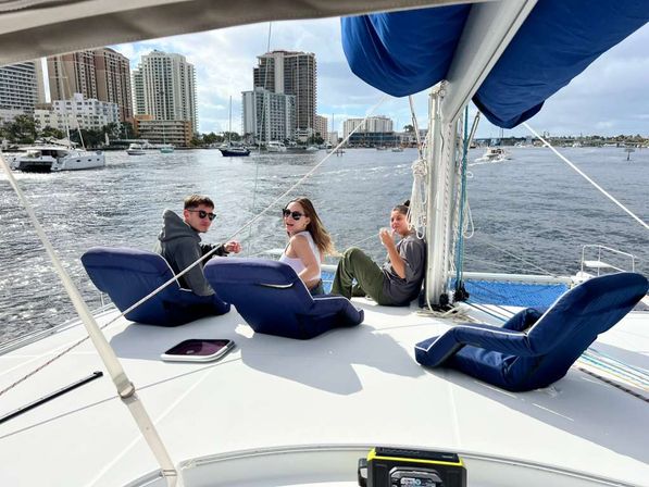 Three people lounging on a sailboat deck with blue cushions and a folded sail, cruising past a marina and coastal city skyline with high-rise waterfront buildings.