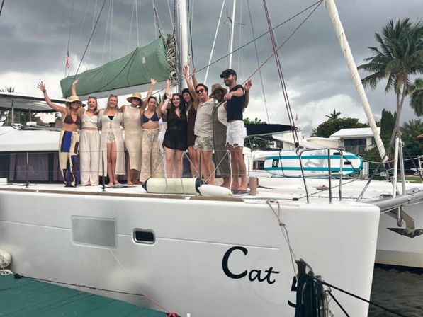 Group of friends waving from the deck of a white catamaran docked at a tropical marina with palm trees and dark storm clouds overhead