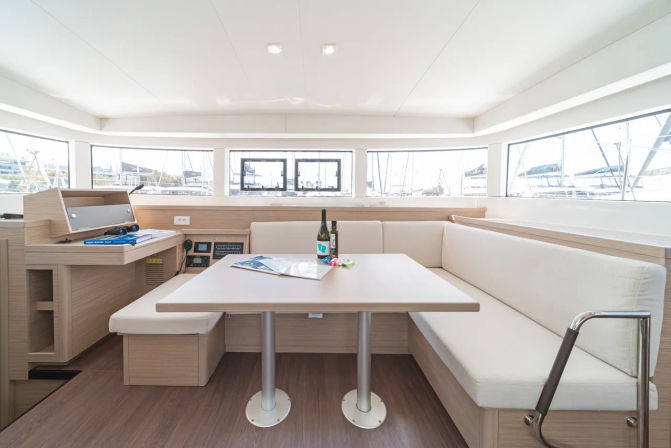 Bright modern yacht salon with light-wood U-shaped dinette and white cushions, central table holding a wine bottle and magazines, panoramic windows revealing marina and sailboats outside.