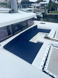 Foredeck lounge on a white catamaran with navy blue cushions, built-in seating and trampoline net, docked at a marina with waterfront homes in the background.