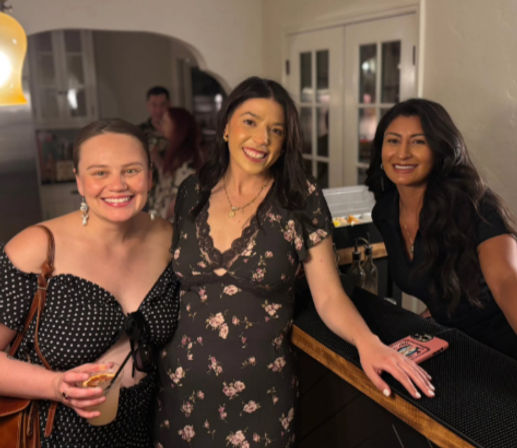 Three smiling women posing at a cozy home kitchen bar during a girls’ night — two holding cocktails, warm lighting and French doors behind them.