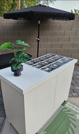 Backyard patio: white outdoor serving station with twelve stainless-steel buffet pans and lids, black fringed umbrella, potted tropical plant and cinderblock wall
