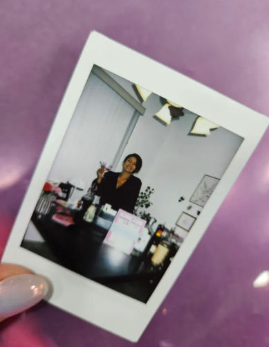 Hand holding a Polaroid of a smiling woman raising a drink behind a home bar setup indoors, with framed art, plants and pendant lights visible against a purple background.