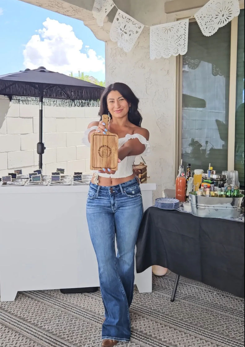 Smiling woman on a sunny backyard patio presenting an engraved wooden cutting board to the camera, wearing an off-shoulder top and jeans, with a drink station and papel picado banners in the background.