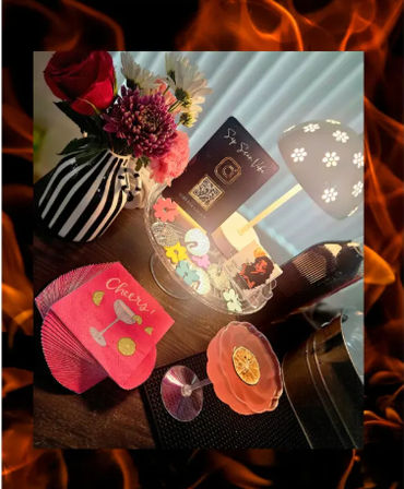 Cozy home-bar tabletop with a pink coupe cocktail garnished with dried orange, bright “Cheers!” napkins, a black-and-white striped vase of mixed flowers, daisy-cut lamp, and a decorative tray with colorful confetti and a QR-code card — playful party setup.