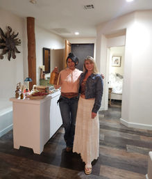 Two smiling women in casual boho outfits pose in a warm, rustic entryway with dark wood floors; one holds a small wooden gift bag beside a white console table topped with snacks, candles and decor, with a bedroom doorway visible in the background.
