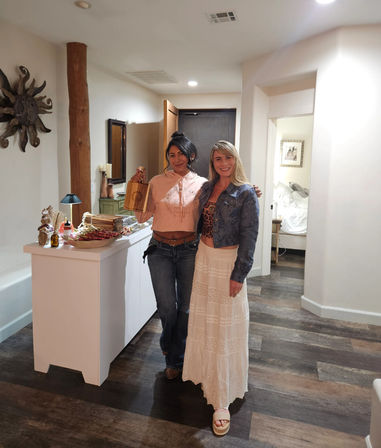 Two smiling women in casual boho outfits pose in a warm, rustic entryway with dark wood floors; one holds a small wooden gift bag beside a white console table topped with snacks, candles and decor, with a bedroom doorway visible in the background.