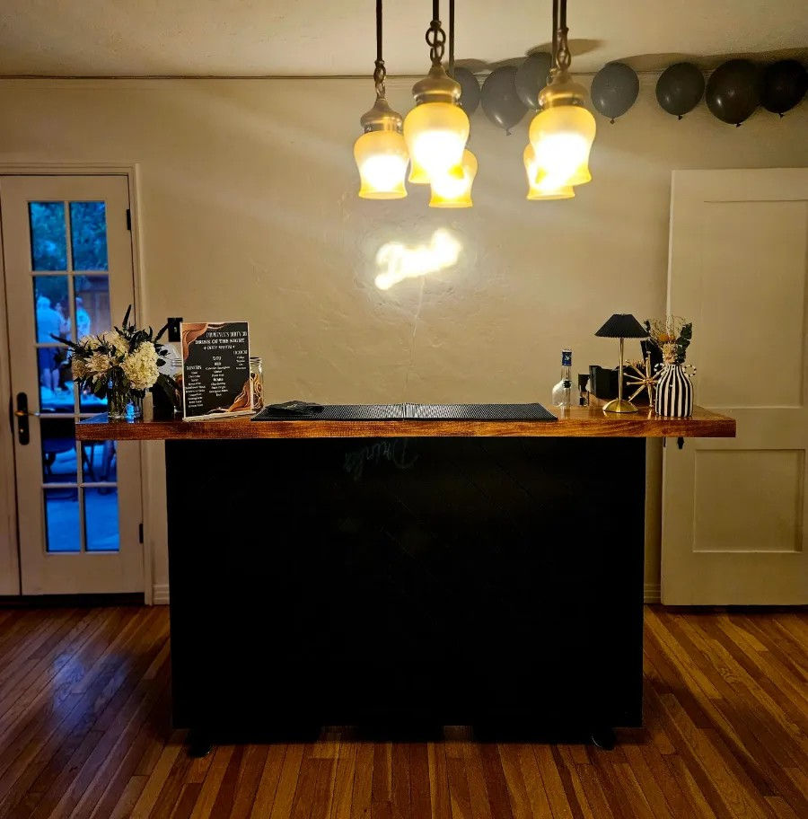 Cozy indoor home bar with a wooden countertop and black base, three warm pendant lights overhead, black balloons along the ceiling, floral arrangements, bottled spirits and a small drink menu on hardwood floors.