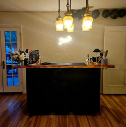 Cozy indoor home bar with a wooden countertop and black base, three warm pendant lights overhead, black balloons along the ceiling, floral arrangements, bottled spirits and a small drink menu on hardwood floors.
