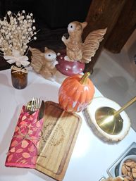 Autumn table centerpiece with playful squirrel figurines on a red mushroom, glossy ceramic pumpkin, dried flower bouquet in an amber vase, wooden serving board with a tied red leaf-pattern napkin and gold cutlery, and a brass lamp on a wood slice.