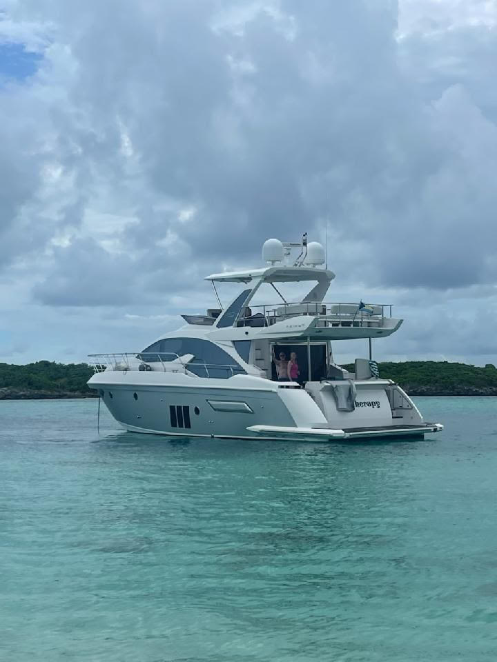 Sleek white luxury motor yacht anchored in clear turquoise tropical water near a green shoreline, cloudy sky overhead, two people on the aft deck.