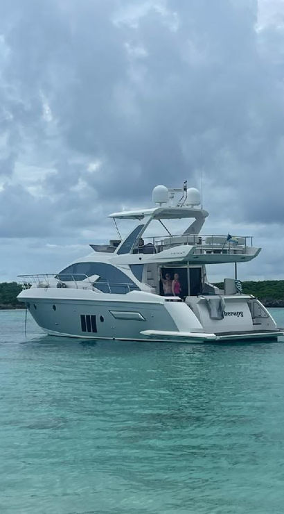 Sleek white luxury motor yacht anchored in clear turquoise tropical water near a green shoreline, cloudy sky overhead, two people on the aft deck.