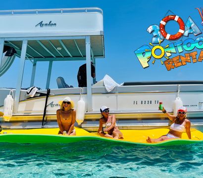 Four women in swimsuits and sun hats lounging and smiling on a bright yellow floating mat beside a white pontoon boat in clear turquoise coastal water on a sunny day, one holding a drink.