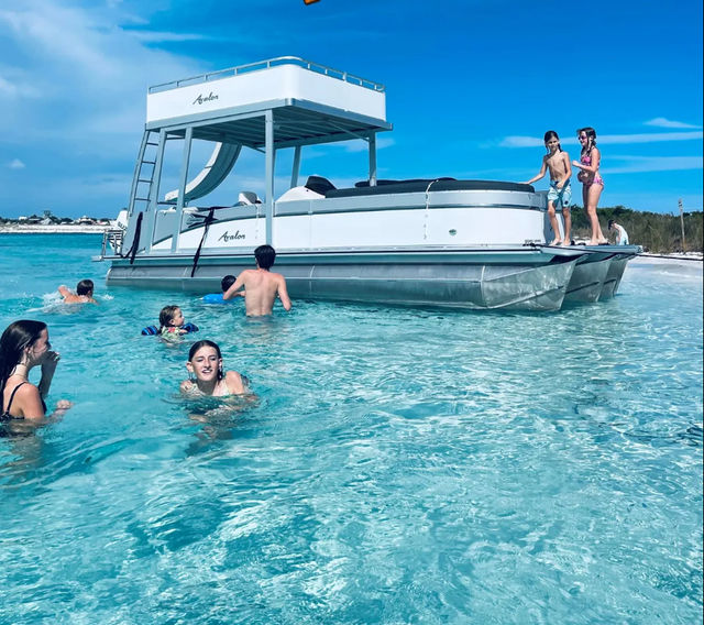 Group of swimmers and kids playing in crystal-clear turquoise shallow water beside a white double-deck pontoon boat anchored near a sandy beach on a sunny day