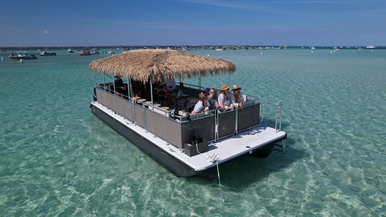Pontoon party boat with a thatched tiki roof and a small group relaxing in clear turquoise shallow bay, anchored among other boats on a sunny day.