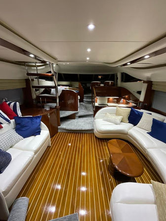 Luxury yacht interior salon with glossy teak plank flooring, curved white leather sofas, navy accent pillows, wood coffee table, stainless ladder and recessed ambient lighting