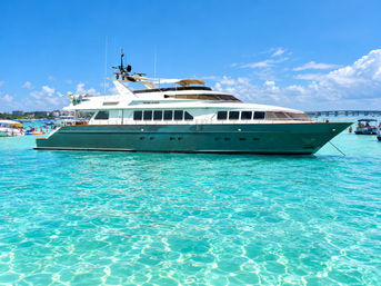 Sleek luxury yacht floating on crystal-clear turquoise water under a bright blue sky, with beaches and other boats visible on the distant shoreline