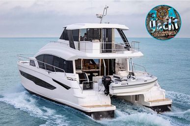 Luxury white motor catamaran cruising calm turquoise ocean, twin-hull wake and rear deck with an inflatable dinghy, modern flybridge and railings.