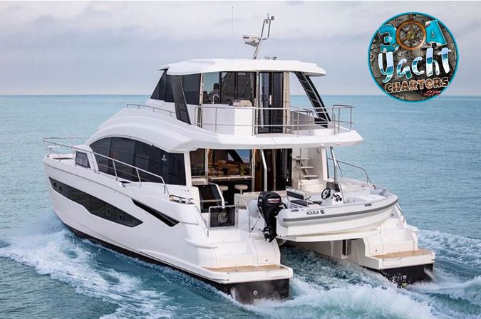 Sleek white luxury catamaran yacht cruising turquoise coastal waters, aft view showing twin hulls, glass cabin, upper deck and inflatable dinghy on the swim platform leaving a frothy wake.