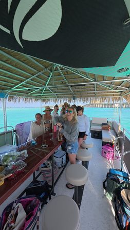 Smiling group on a thatched-roof pontoon boat opening a bottle and enjoying turquoise coastal waters by a pier on a sunny day