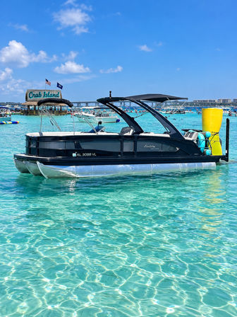 Black pontoon boat anchored in crystal-clear turquoise water beside a busy sandbar with a thatched floating bar and flags on a sunny blue-sky day