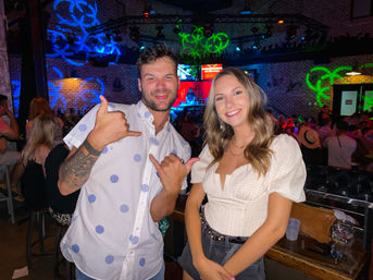 Smiling young man and woman posing inside a lively nightclub bar with exposed brick walls, colorful neon stage lights, dancing crowd in the background, and the man flashing a shaka hand sign.