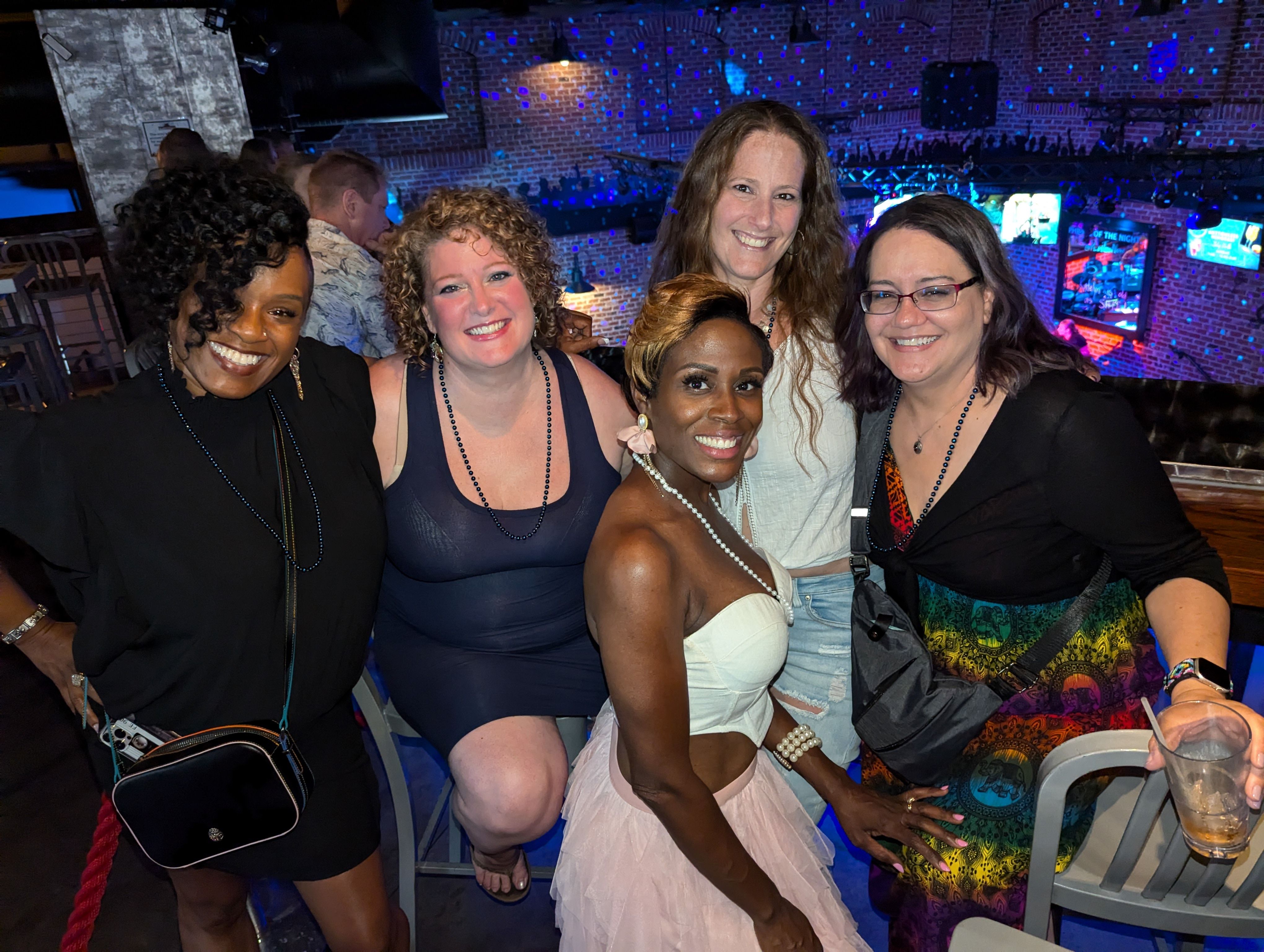 Five women smiling and posing together at a lively brick‑walled nightclub bar with colorful disco lights and TV screens in the background.