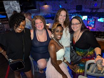 Five women smiling and posing together at a lively brick‑walled nightclub bar with colorful disco lights and TV screens in the background.