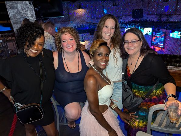 Five women smiling and posing together at a lively brick‑walled nightclub bar with colorful disco lights and TV screens in the background.