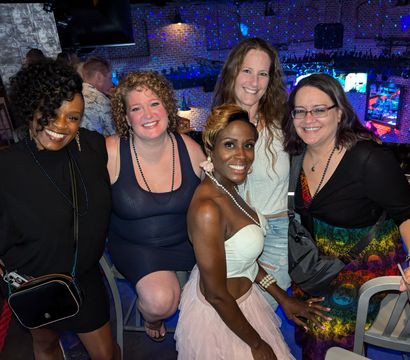 Five women smiling and posing together at a lively brick‑walled nightclub bar with colorful disco lights and TV screens in the background.